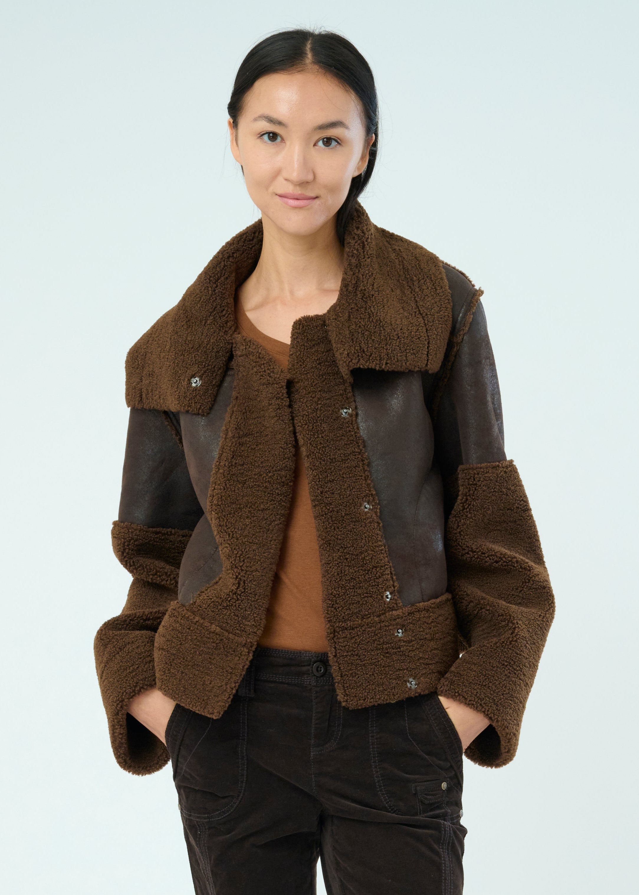 Front view of a tall female model with long black hair confidently posing wearing our dark chocolate colored Trisha Bonded Distressed Suede Sherpa Jacket from Marrakech Clothing.